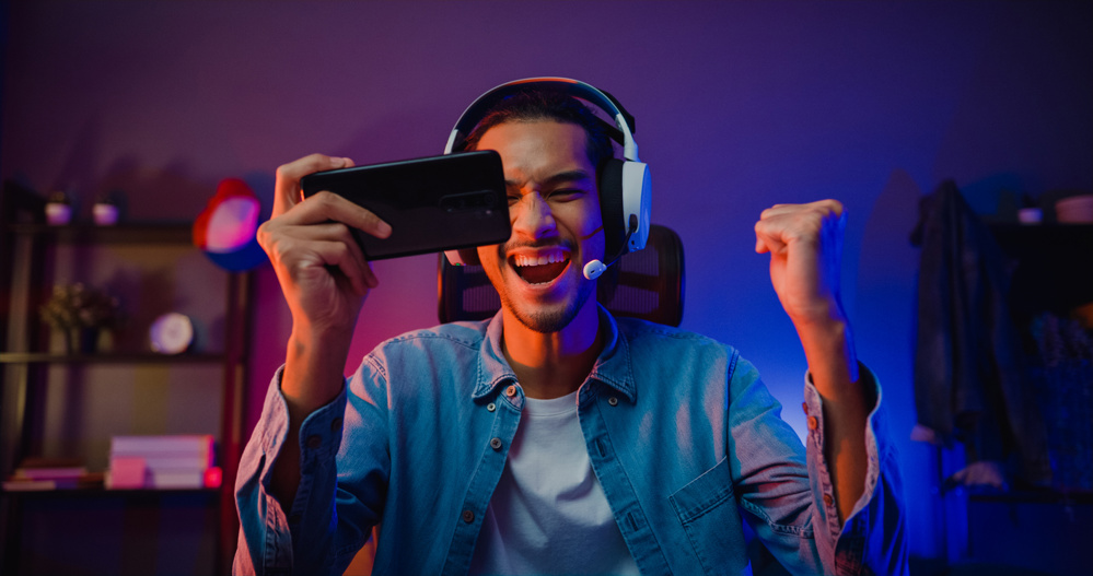 Happy Asia man gamer wear headphone competition play video game online with smartphone colorful neon light in living room at night modern house. Esport streaming game online, home quarantine activity